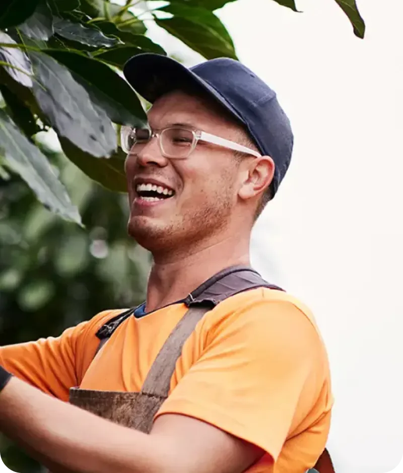 A smiling horticulture trainee in a cap and safety glasses, working among plants