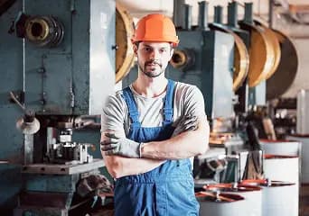 A machine operator in overalls and a hard hat beside a press