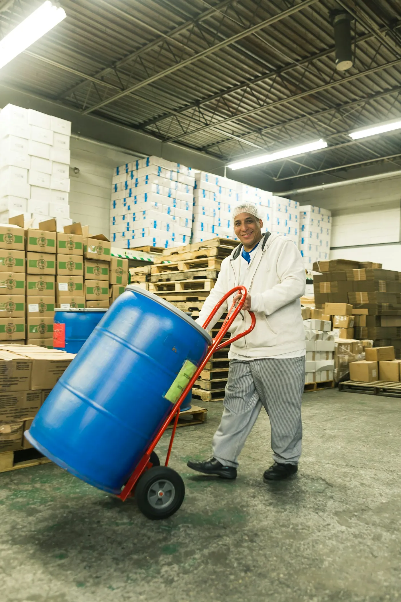 A smiling warehouse worker moving a blue drum on a hand trolley